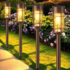 four lights that are on the side of a sidewalk in front of some bushes and flowers