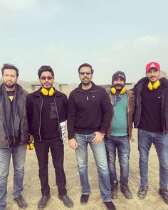 four men standing in front of a fence with ear phones on their ears and one wearing a black jacket