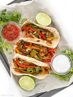 three tacos with peppers, onions and cilantro on parchment paper next to salsa