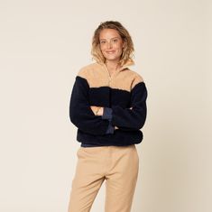 a woman standing in front of a white wall wearing a black and tan sweater with her arms crossed