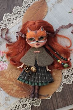 a doll with long red hair wearing a green dress and orange mask on top of a lace doily