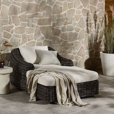 a wicker chaise lounge chair sitting in front of a stone wall with potted plants