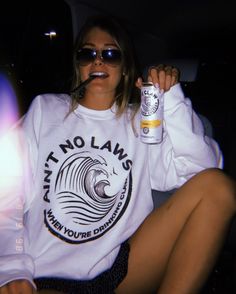 a woman sitting in the back of a car holding a can of beer and wearing sunglasses