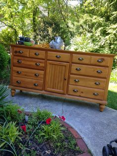 a wooden dresser sitting on top of a cement ground next to flowers and trees in the background