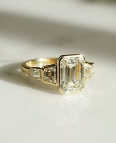 an emerald cut diamond ring with three baguets on the side, sitting on a white surface