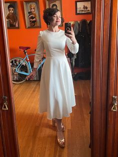 a woman in a white dress taking a selfie with her cell phone while standing in front of an orange wall