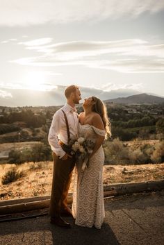 a man and woman standing next to each other on top of a hill with mountains in the background