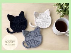 three crocheted cat coasters sitting on top of a table next to a cup of coffee