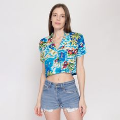 Vintage 80s cropped button up blouse in blue with a Hawaiian-themed print of palm trees, volcanos, and flowers. Measurements and Condition:    Fits like: Labeled women's medium Fabric: Rayon - labeled machine wash cold Brand: Andrew Sport, made in USA Condition: Excellent, appears to have been hemmed up by a previous owner.   Length: 15.5" Chest: 39" Waist: 38" Shoulders (seam to seam): 16" Sleeve: 6.5" Shown on a 5'9" model with measurements of 33"-24"-37", usually wears size XS to small. See our FAQ for more info on sizing and condition ratings. Tropical Vacation Tops With Camp Collar, Tropical Tops With Camp Collar For Vacation, Blue Hawaiian Style Summer Tops, Blue Hawaiian Summer Top, Tropical Camp Collar Tops For Vacation, Blue Hawaiian Shirt With Hibiscus Print For Summer, Blue Hawaiian Shirt With Hibiscus Print For Beach, Blue Hawaiian Beach Top, Blue Tropical Print Hawaiian Shirt For Vacation