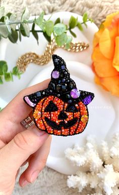 a hand holding a beaded halloween pumpkin brooch on a white plate with flowers in the background