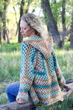 a woman sitting on a wooden bench wearing a crocheted sweater with an open hood