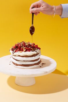 a person drizzling chocolate onto a cake on a white plate with yellow background