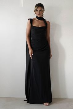 a woman in a long black dress standing next to a white wall with her hands on her hips