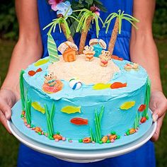 a person holding a blue cake decorated with animals and palm trees on the top, in front of some flowers