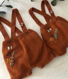 two brown knitted baby overalls with flowers and leaves on them, sitting on a white sheet