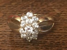 This gorgeous ring features a sparkling cluster of cubic zirconia stones, paired with lustrous 14 karat yellow gold. The band is an approximate size 6.5, and the ring weighs approximately 4.5 grams. This gorgeous piece has a unique tiered cluster of stones at center that adds to its appeal and intrigue. In addition, the shoulders of the band have a unique, heart or leaf shaped detail. This is the ideal piece to buy for yourself, and it would also make a beautiful promise ring, graduation gift, a Yellow Gold Cluster Ring With Cubic Zirconia, Dazzling Gold Cluster Diamond Ring, Gold Cluster Diamond Ring With Accents, 14k Gold Cluster Ring With Prong Setting, Yellow Gold Diamond Cluster Ring, Yellow Gold Cluster Ring For Promise, Dazzling Gold Cluster Ring With Center Stone, Sparkling Cluster Diamond Ring, Dazzling White 14k Gold Cluster Ring