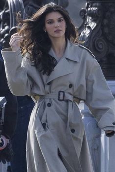 a woman in a trench coat walking down the street with her hand on her hip