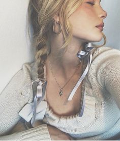 a woman with blonde hair wearing a white sweater and silver bow tie necklace, leaning against a wall