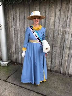 a woman in a blue dress and hat standing next to a fence