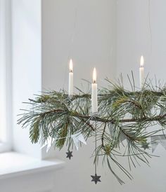 three lit candles are hanging from a pine branch with silver stars on the branches and white background