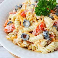 a pasta salad with tomatoes, olives and blueberries in a white bowl on a wooden table