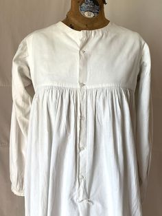 "1815. 32\" bust, white cotton chemise  with long wide sleeves, round neckline, bounle lined yoke, high waisted bodice. Chemise has full skirt to floor and fastens in front, from neck to below knees  with 5 small white glass buttons. Measurements: 32\" bust  Shoulder to shoulder 14\" Shoulder to hem  46\" Ar.holes 7\" Sleeves 20\" Neckline 7\" wide, 3\" deep  Width at bottom 72\" Condition very good,  small rust dots in front" White Cotton Regency Prairie Dress, White Cotton Regency Style Prairie Dress, Victorian Cotton Prairie Dress With Long Sleeves, Long Sleeve Cotton Dresses With Historical Design, Long Sleeve Cotton Dress With Historical Design, Cotton Victorian Dress With Buttons For Daywear, Cotton Long Sleeve Dresses With Historical Design, Classic Cotton Prairie Dress For Daywear, Cotton Victorian Dress With Long Sleeves In Regency Style