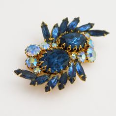 Stunning blue rhinestone brooch incorporates blue navette tones, ab blue faceted chatons, and ovel faceted blue stones which have a fancy prong setting. The colors are striking and bold in the gorgeous unsigned mid-century brooch. It measures 2 1/2 inches side to side and 1 1/2 inches top to bottom. It is in very good vintage condition with some very light wear to the ab coating on a couple of stones - nothing noticeable when wearing. A wonderful statement piece on a white dress for spring! Vintage Rhinestone Jewelry, Mid Century Jewelry, Blue Stones, Dress For Spring, Floral Accessories, Brooch Vintage, Floral Jewellery, Rhinestone Jewelry, Rhinestone Brooches