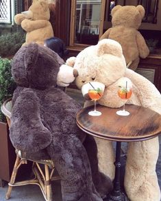 two large teddy bears sitting at a table with drinks in front of them on the sidewalk