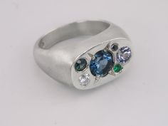 Journey back to an era of opulence with this candy studded cushion signet ring. At its centre, a mesmerising 6mm London blue topaz captures the heart of celestial beauty, surrounded by a constellation of gems: 4mm lilac grey spinel, 3mm Australian teal sapphire, 3mm moissanite, 2mm lab emerald, and a 1.9mm salt and pepper diamond. Each gem is thoughtfully chosen to create a harmonious blend of colours and light, echoing the ancient world's fascination with the cosmos. This ring not only celebrat Celestial Oval Multi-stone Ring, Celestial Oval Signet Ring For Anniversary, Teal Sapphire, Lilac Grey, Pepper Diamond, Pearl Gemstone, Black Rhodium, Salt And Pepper Diamond, Raw Gemstones