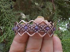 a hand holding a piece of wire with purple beads on it's end and some branches in the background