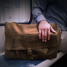 a man's hand is on top of a brown leather messenger bag in an airplane