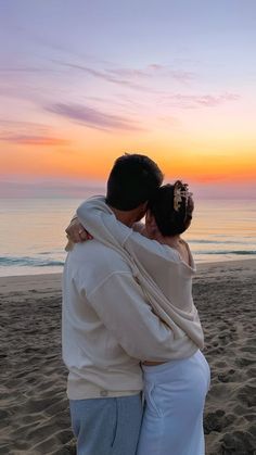 a man and woman hug on the beach as the sun sets in the distance behind them