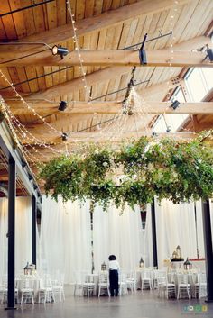 an indoor wedding venue with white chairs and greenery hanging from the ceiling over tables