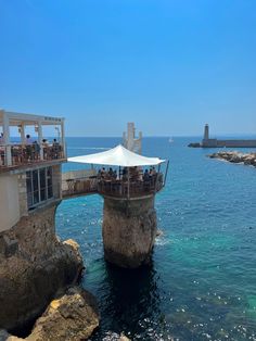 people are sitting at tables on the rocks by the water and under an umbrella over looking the ocean