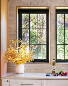 a vase with flowers and fruit sitting on a counter in front of two large windows