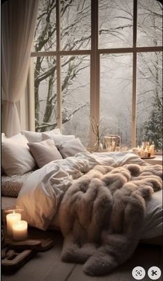 a bed with blankets, pillows and candles in front of a window that has snow outside