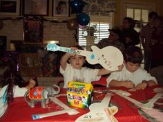 three children sitting at a table with toys and balloons in the background as people look on