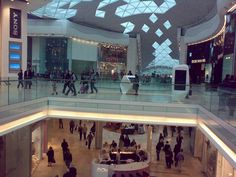 people are walking around in an indoor shopping mall with lights on the ceiling and large glass windows