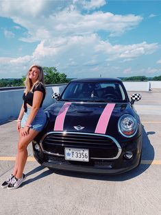 a woman sitting on the hood of a black mini cooper with pink and white stripes