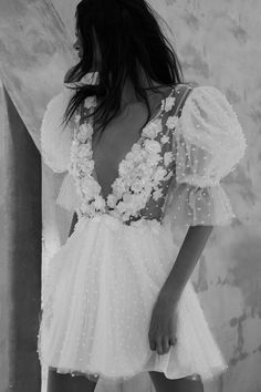 a woman in a short white dress with flowers on her shoulders and back, standing next to a wall