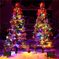 two christmas trees decorated with multicolored lights in the middle of snow covered ground