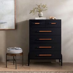 a black chest of drawers next to a small stool with a plant on top and a painting hanging above it