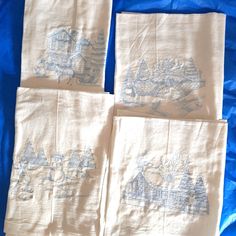 four embroidered tea towels with houses and trees in blue ink on white linen, laid out on a blue sheet