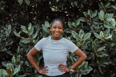 a woman standing with her hands on her hips in front of some bushes and trees