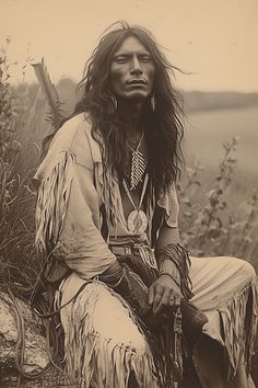 an old photo of a native american man with long hair and feathers sitting on a rock