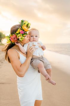 a woman holding a baby wearing a flower crown on her head while standing on the beach