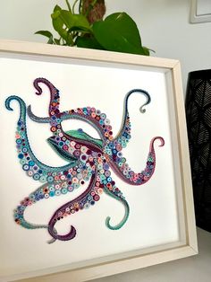 an octopus made out of buttons is displayed in a white frame next to a potted plant