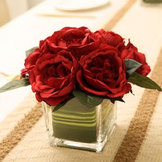 red roses in a square glass vase on a dining room table with place mats and napkins