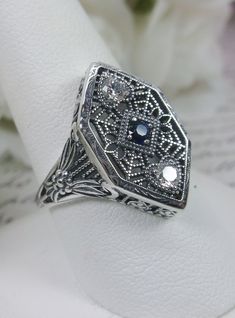 Blue Sapphire & White Cubic Zirconia (CZ) Sterling Silver RingCharlotte Design#D231 Resurrected from the Art Deco movement, this gorgeous Art Deco reproduction filigree ring is crafted in sterling silver. This lovely filigree ring is set with a stunning quality 2mm center round-cut simulated blue sapphire gemstone. Two small white cubic zirconia gemstones also grace the face of the ring, one near each point, each set in the center of a tiny heart. There are 2 filigree accent flowers that grace t Antique Silver Sapphire Ring With Diamond Accents, Victorian Filigree Ring With Intricate Design In Sterling Silver, Sterling Silver Victorian Filigree Ring, Victorian Sterling Silver Filigree Ring With Intricate Design, Vintage Cubic Zirconia Filigree Ring With Intricate Design, Vintage Filigree Ring With Intricate Design In Cubic Zirconia, Silver Sapphire Ring With Intricate Design, Ornate Filigree Ring For Anniversary, Stamped 925, Ornate Sterling Silver Filigree Ring With Intricate Design