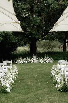 an outdoor ceremony setup with white chairs and flowers on the grass, under umbrellas